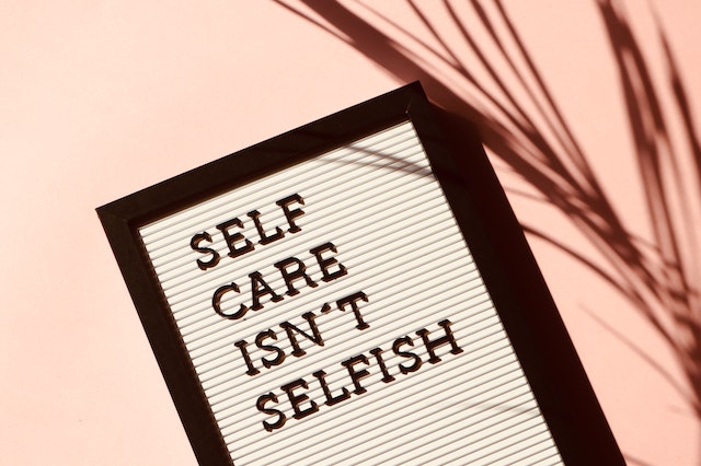 Self Care Isn't Selfish written on a board with an out of focus plant in the foreground