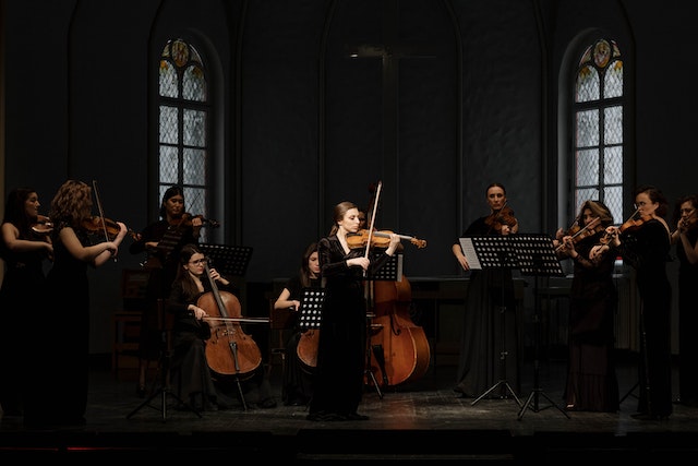 A woman wearing a black dress, standing and playing a violin in a string ensemble with 10 other musicians in a darkly lit corner of a church